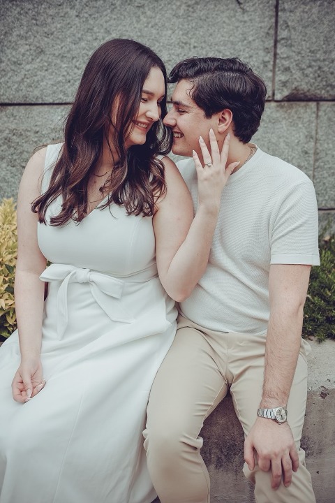 beautiful engaged couple dressed in white, seated in front of a stone wall